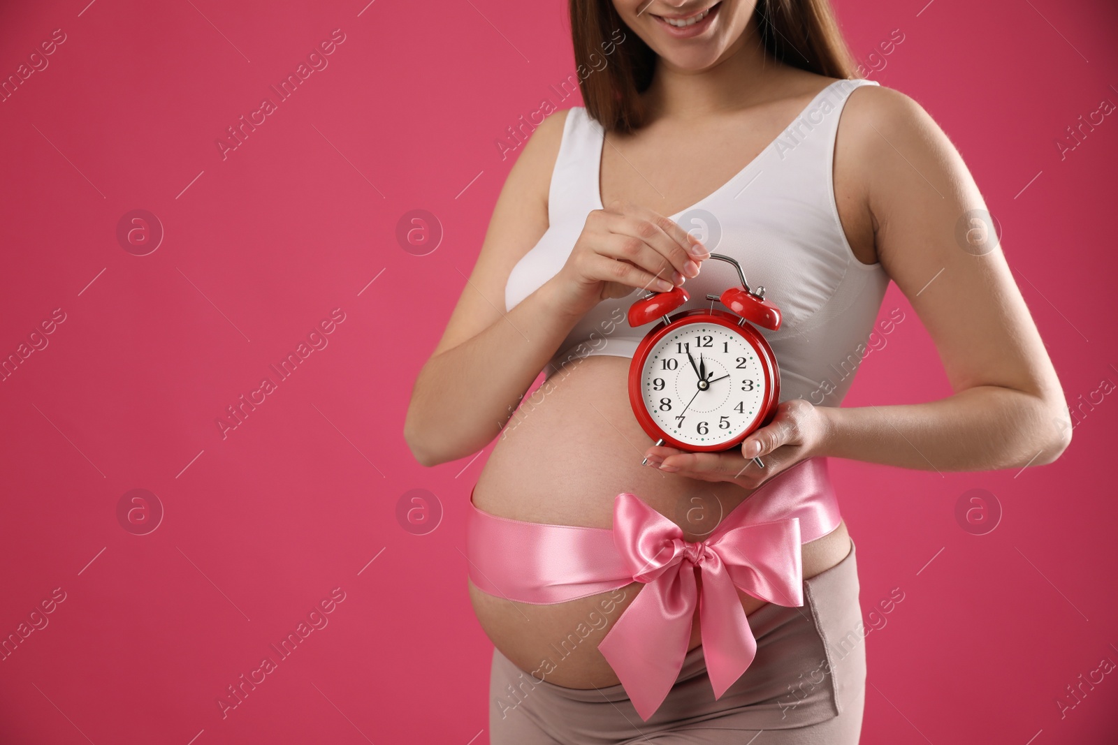 Photo of Young pregnant woman with alarm clock and bow on pink background, closeup. Time to give birth