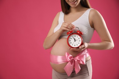 Young pregnant woman with alarm clock and bow on pink background, closeup. Time to give birth