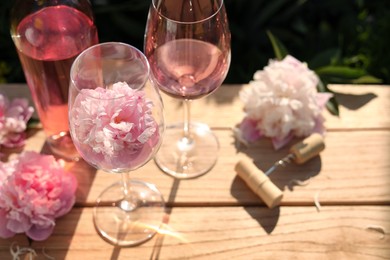 Photo of Bottle and glasses of rose wine near beautiful peonies on wooden table outdoors
