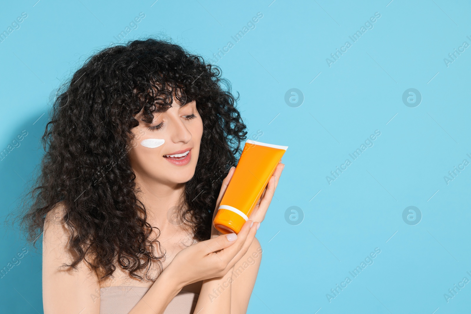 Photo of Beautiful young woman with sun protection cream on her face holding tube of sunscreen against light blue background, space for text