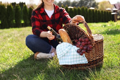 Photo of Woman taking bottle of wine from wicker basket outdoors, closeup. Picnic season