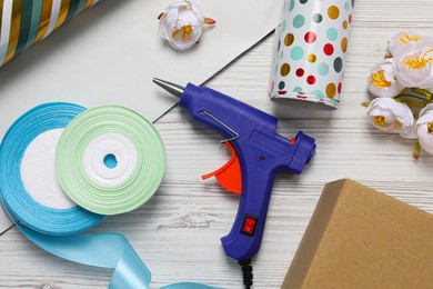 Photo of Hot glue gun and handicraft materials on white wooden table, flat lay