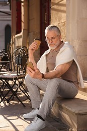 Handsome senior man sitting on doorstep, using smartphone and drinking coffee outdoors