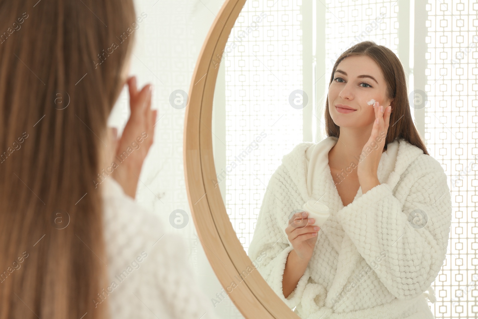 Photo of Young woman applying face cream onto her face near mirror in bathroom