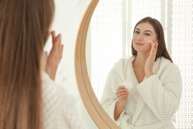 Photo of Young woman applying face cream onto her face near mirror in bathroom