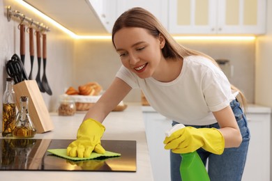 Woman with spray bottle and microfiber cloth cleaning electric stove in kitchen