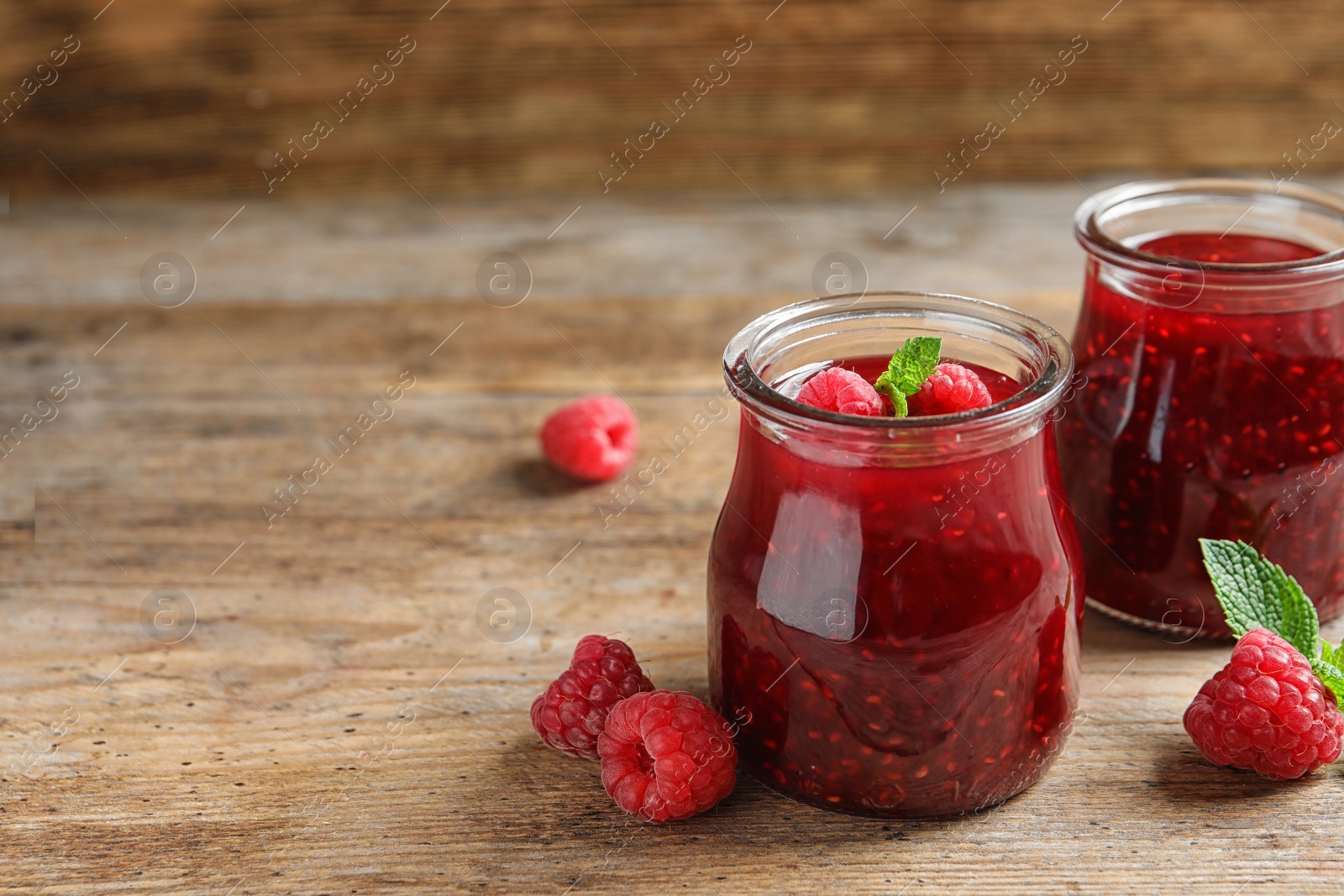 Photo of Glass jars of sweet jam with ripe raspberries and mint on wooden table. Space for text