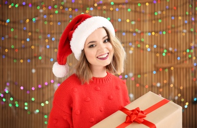 Photo of Beautiful young woman in Santa hat with Christmas gift box on blurred lights background