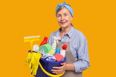 Happy housewife holding bucket with cleaning supplies on orange background