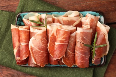 Photo of Rolled slices of delicious jamon with rosemary on wooden table, top view