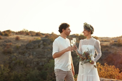 Photo of Happy newlyweds with beautiful field bouquet and glasses of champagne outdoors