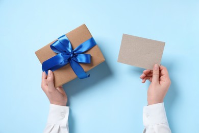 Woman holding gift box and blank card on light blue background, top view. Space for text