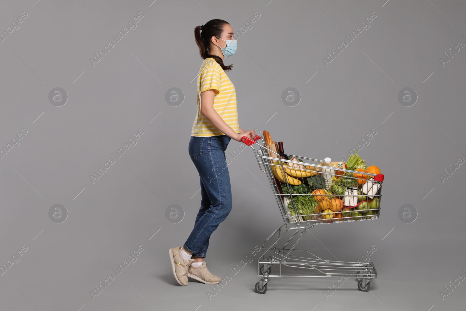 Photo of Woman with protective mask and shopping cart full of groceries on light grey background