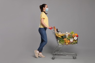 Photo of Woman with protective mask and shopping cart full of groceries on light grey background