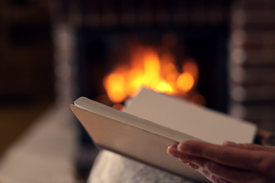 Woman reading book near burning fireplace at home, closeup