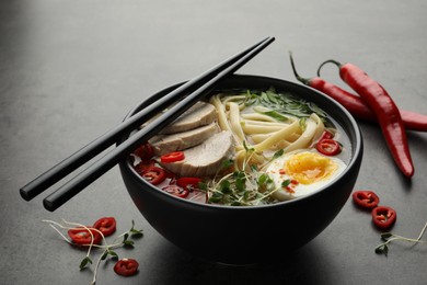 Photo of Delicious ramen with meat in bowl, chopsticks and chili peppers on grey textured table. Noodle soup