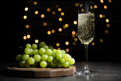 Photo of Green grape and glass of champagne on grey table against black background with blurred lights. Bokeh effect