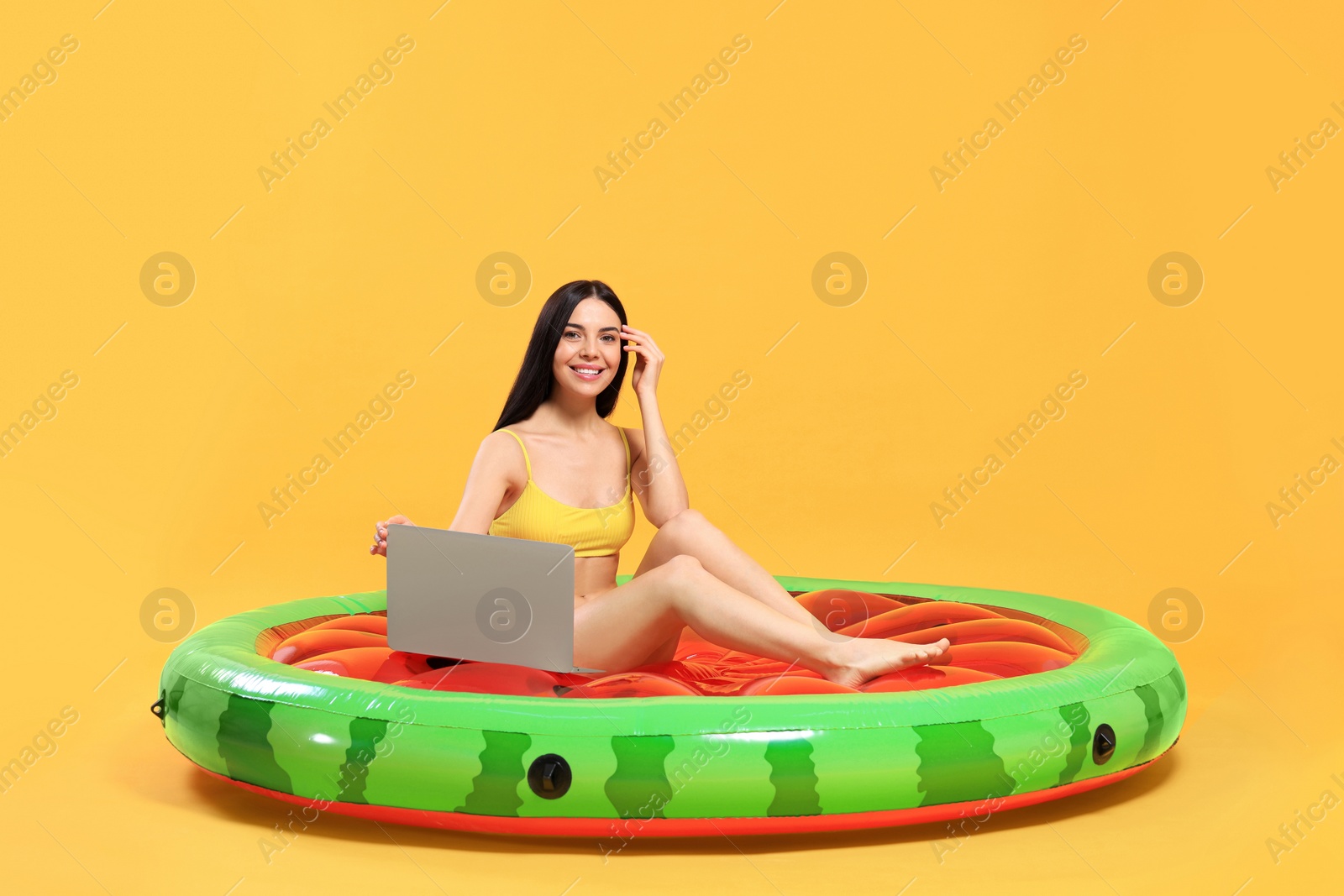 Photo of Happy young woman with beautiful suntan using laptop on inflatable mattress against orange background
