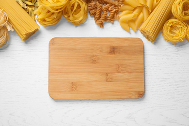 Photo of Different types of pasta and cutting board on white wooden table, flat lay. Space for text