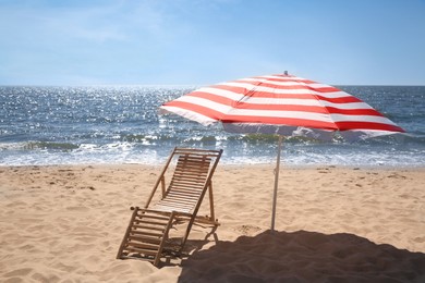 Photo of Deck chair near red and white striped beach umbrella on sandy seashore