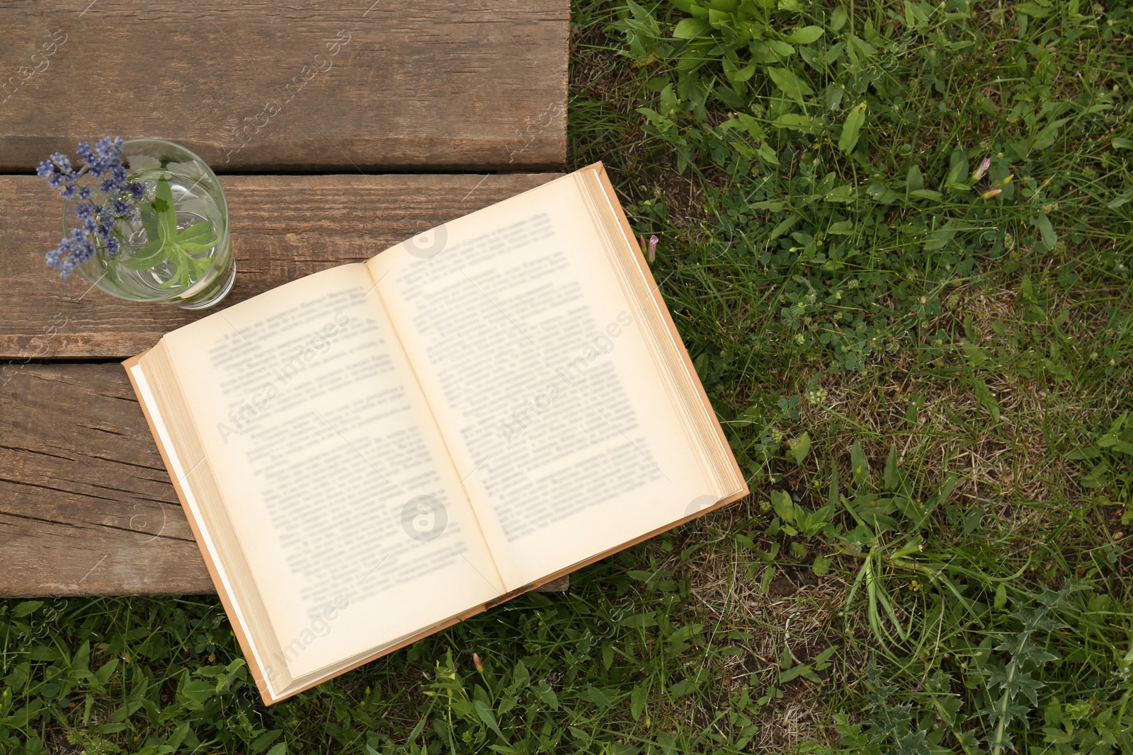 Photo of Open book with flowers in glass on green grass outdoors, top view. Space for text