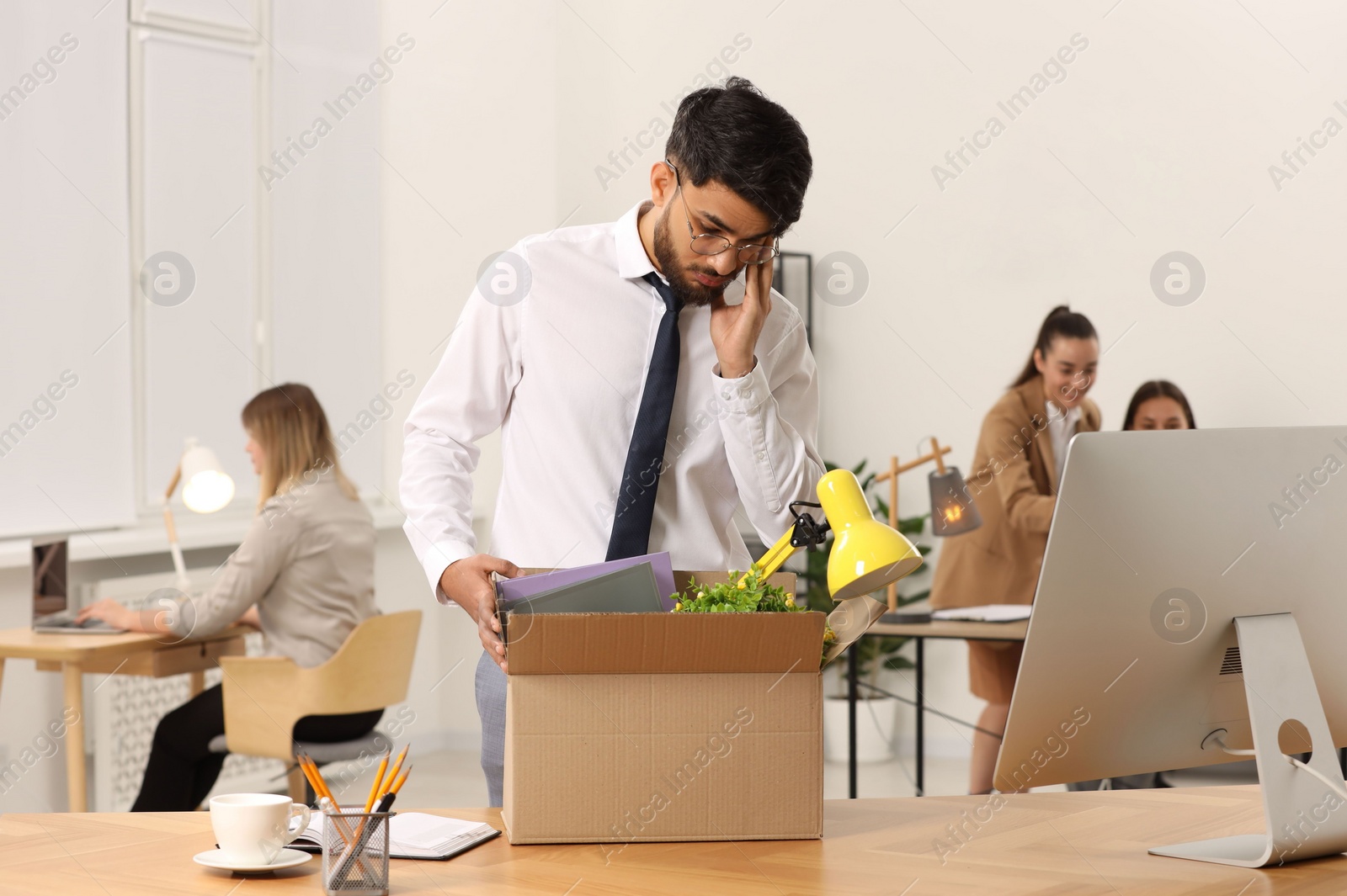 Photo of Unemployment problem. Frustrated man with box of personal belongings at table in office