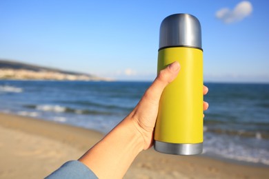 Woman holding yellow thermos with hot drink on beach near sea, closeup. Space for text