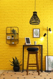 High wooden table with tablet and stool as stand up workplace near yellow brick wall. Stylish interior