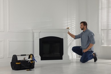 Photo of Man using construction level for installing electric fireplace near white wall in room