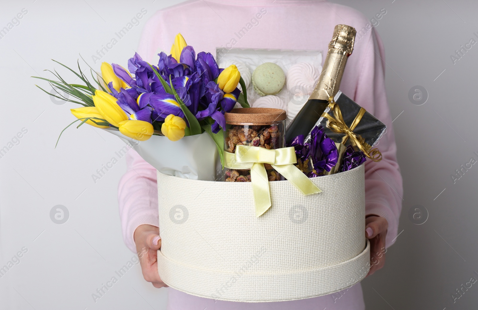 Photo of Woman holding basket with gifts, bouquet and champagne on light grey background, closeup