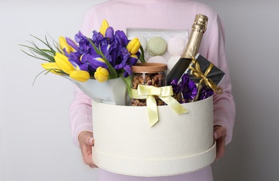 Woman holding basket with gifts, bouquet and champagne on light grey background, closeup