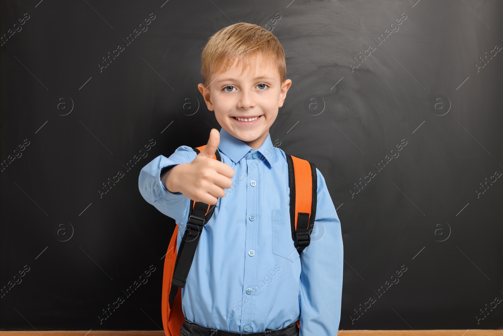 Photo of Happy schoolboy with backpack showing thumb up gesture near blackboard