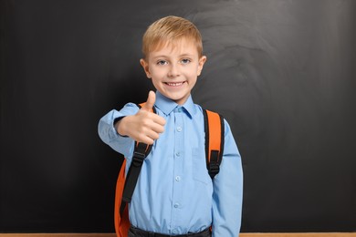 Happy schoolboy with backpack showing thumb up gesture near blackboard