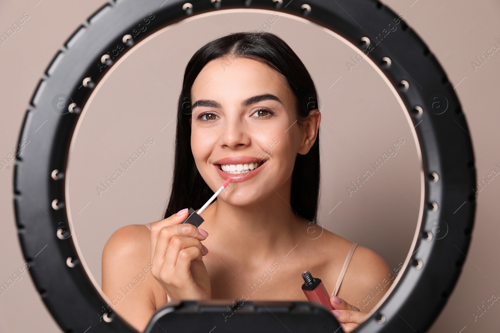 Photo of Beautiful young woman applying liquid lipstick on beige background, view through ring lamp