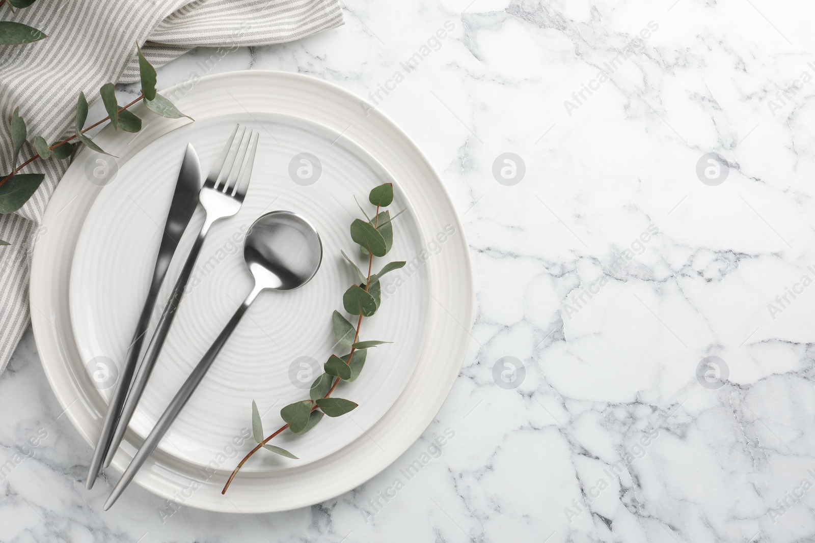 Photo of Stylish setting with cutlery, napkin, eucalyptus branches and plates on white marble table, top view. Space for text