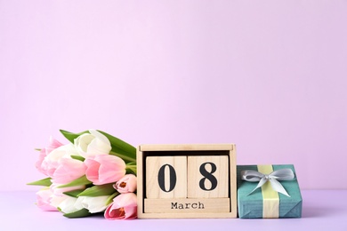 Photo of Wooden block calendar with date 8th of March, tulips and gift box on lilac background, space for text. International Women's Day