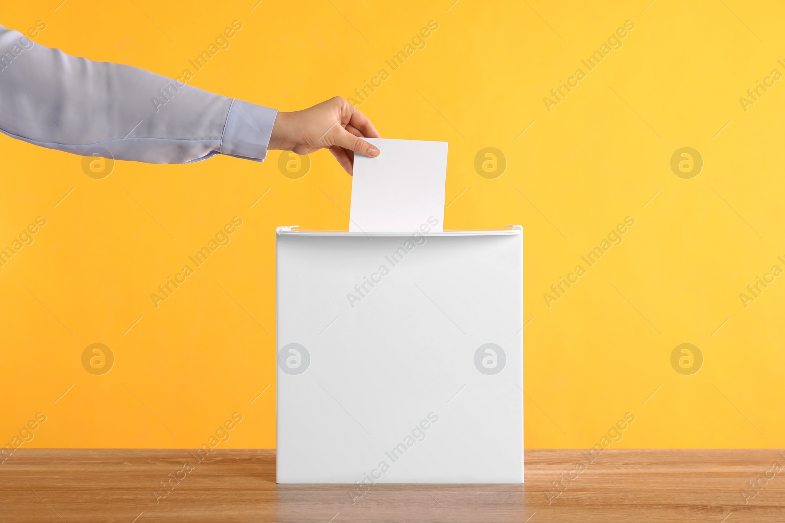 Photo of Woman putting her vote into ballot box on wooden table against orange background, closeup