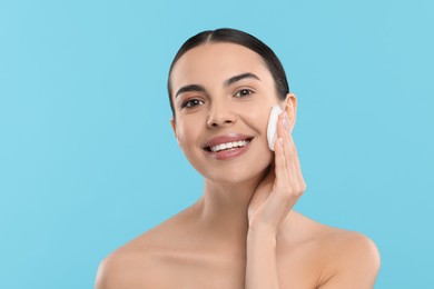 Beautiful woman removing makeup with cotton pad on light blue background