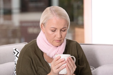 Photo of Ill mature woman with cup of hot tea for cough on sofa at home