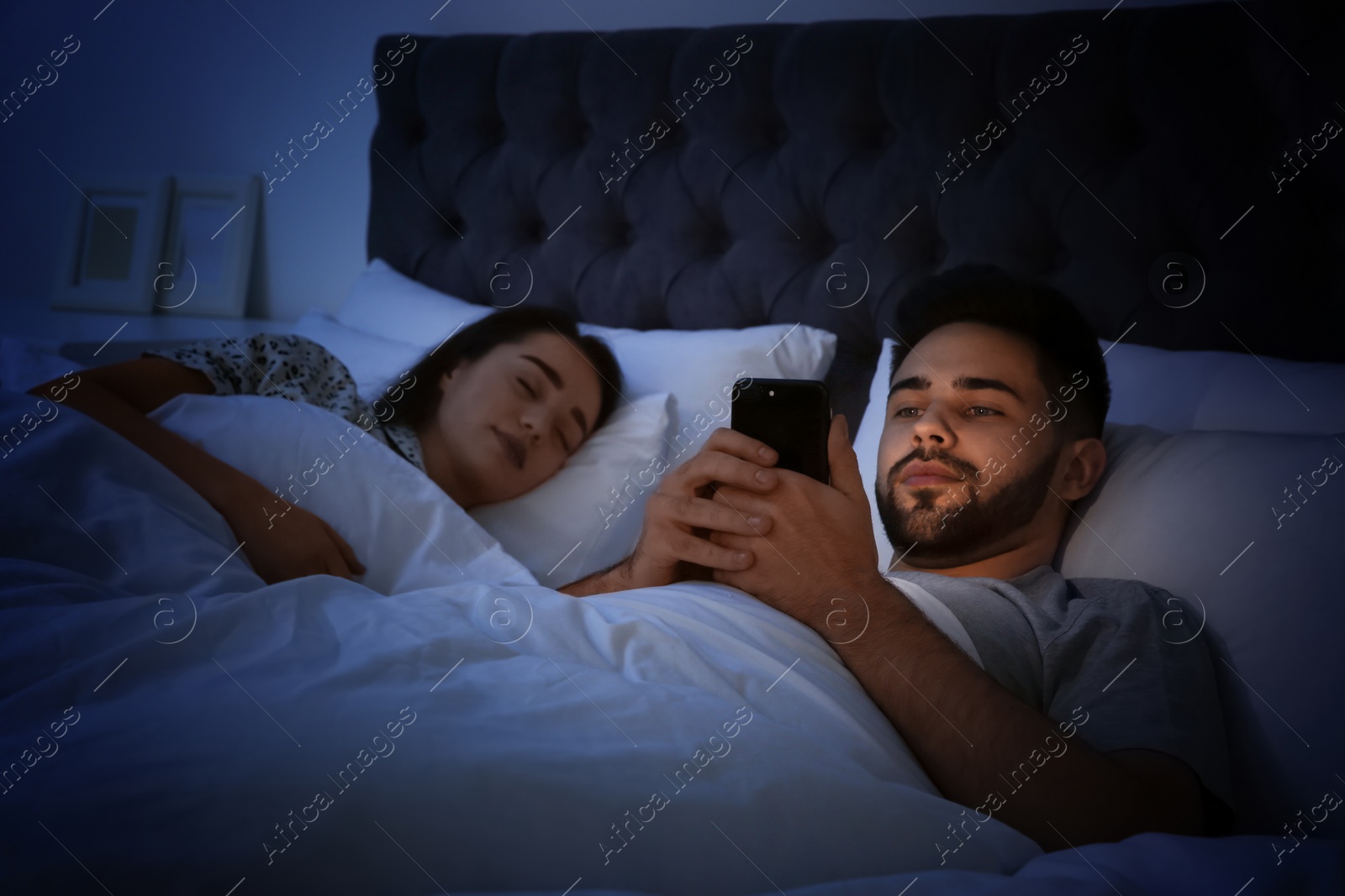 Photo of Young man using smartphone while his girlfriend sleeping in bed at night