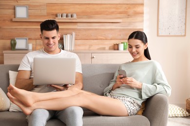 Happy couple in pyjamas with gadgets spending time together on sofa at home