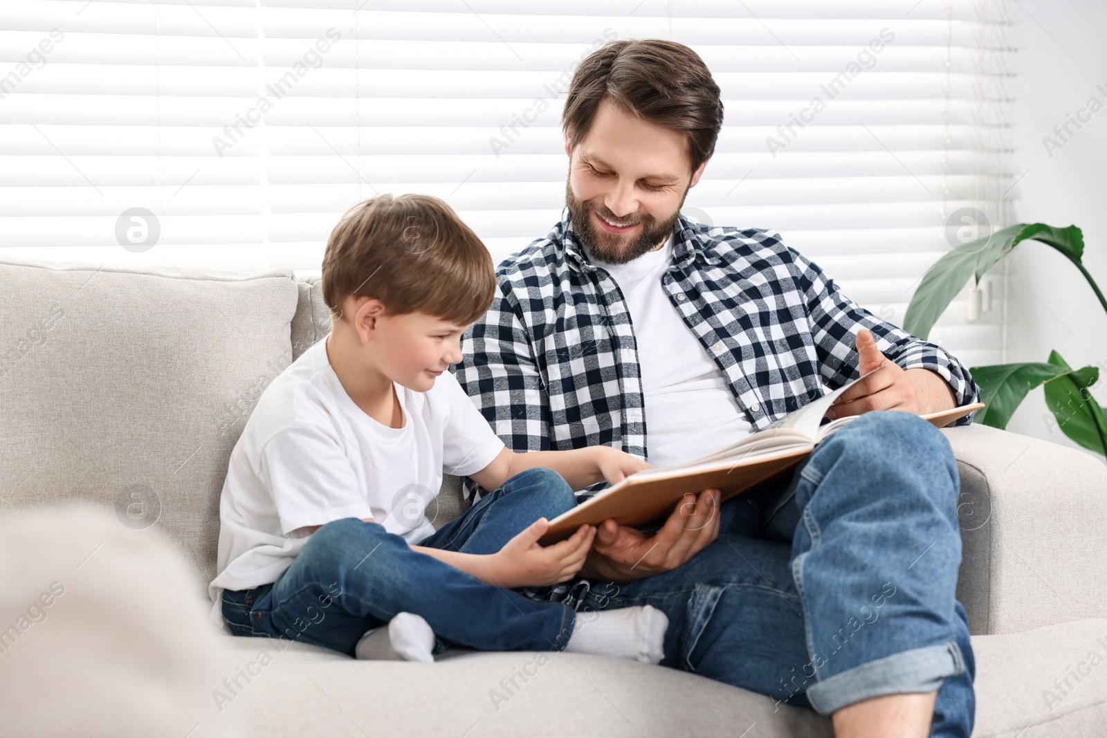 Photo of Happy dad and son reading book together on sofa at home