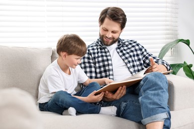 Photo of Happy dad and son reading book together on sofa at home