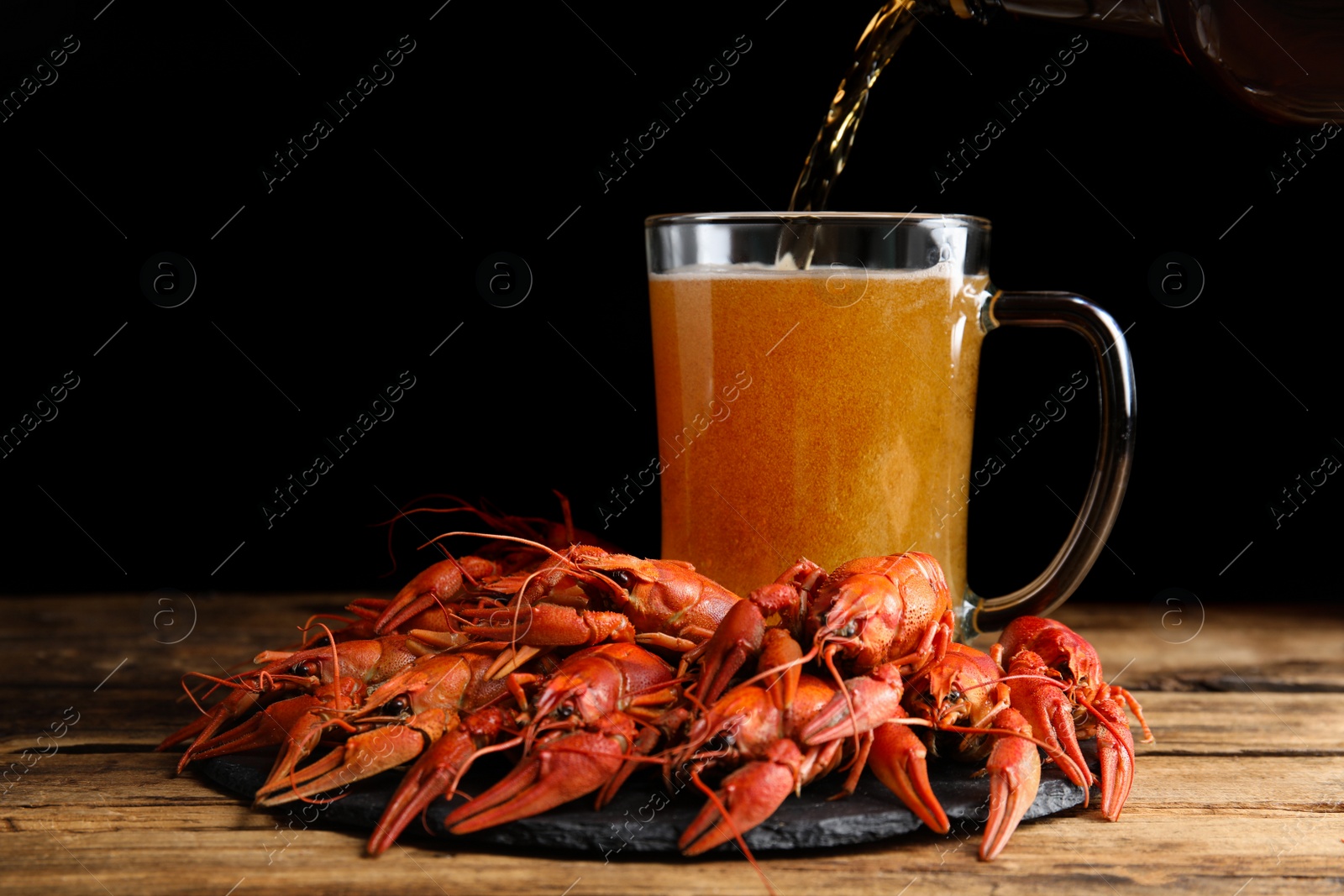 Photo of Delicious red boiled crayfishes and pouring beer into mug on wooden table