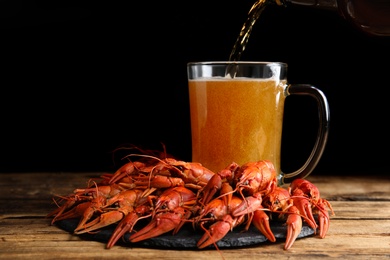 Photo of Delicious red boiled crayfishes and pouring beer into mug on wooden table