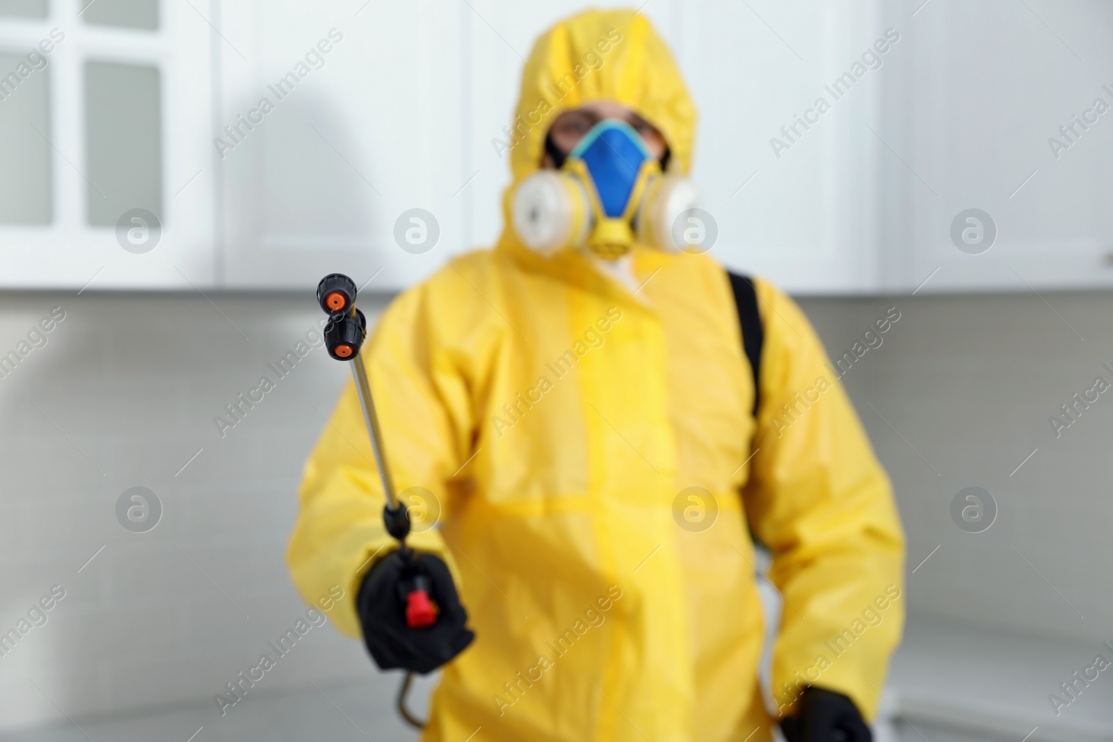 Photo of Pest control worker in protective suit indoors, focus on insecticide sprayer