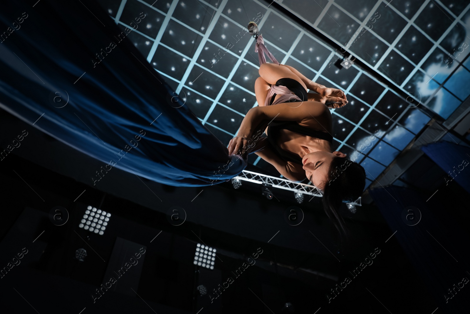 Photo of Young woman performing acrobatic element on aerial silk indoors, low angle view