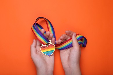 Photo of Woman holding rainbow ribbon with heart shaped pendant on orange background, top view. LGBT pride