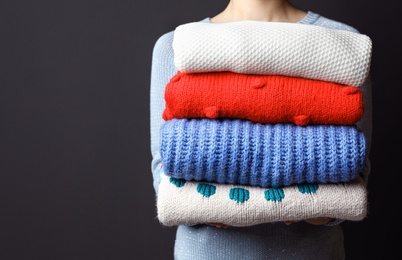 Woman holding pile of winter sweaters on dark background, closeup view. Space for text