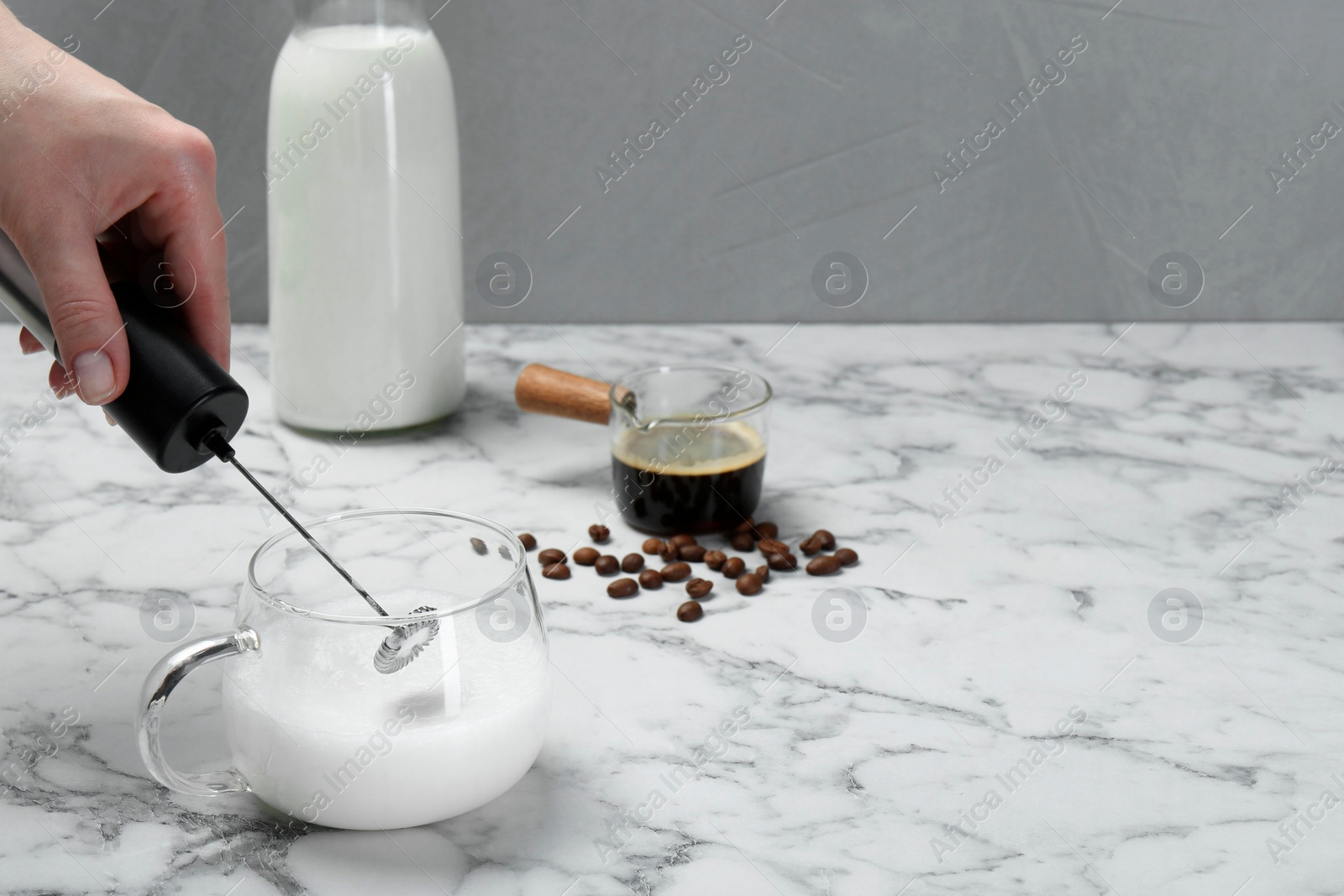 Photo of Woman whisking milk in cup with mini mixer (milk frother) at white marble table, closeup. Space for text